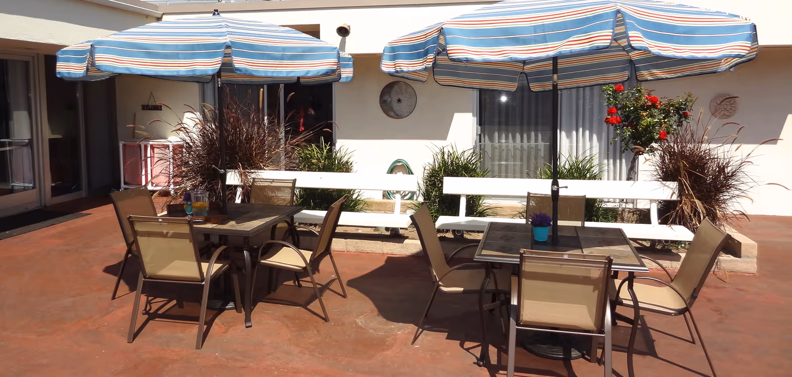 Outdoor patio area with two tables, each surrounded by four chairs and shaded by blue and white striped umbrellas. There are white benches and plants along the wall of the building in the background.