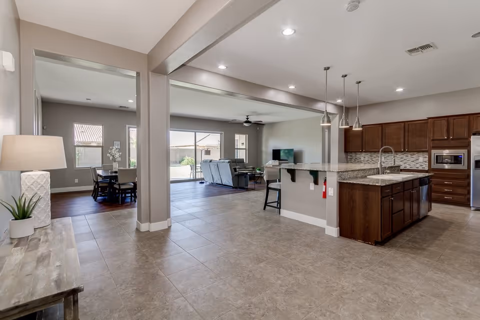 Open-concept interior space featuring a kitchen with dark wood cabinets, granite countertops, and pendant lighting over a breakfast bar with stools. Adjacent living room area with reclining sofas and a TV, and a dining area with a table and chairs near windows letting in natural light. Neutral color walls and tiled flooring in the kitchen and living area, with wood flooring in the dining space.