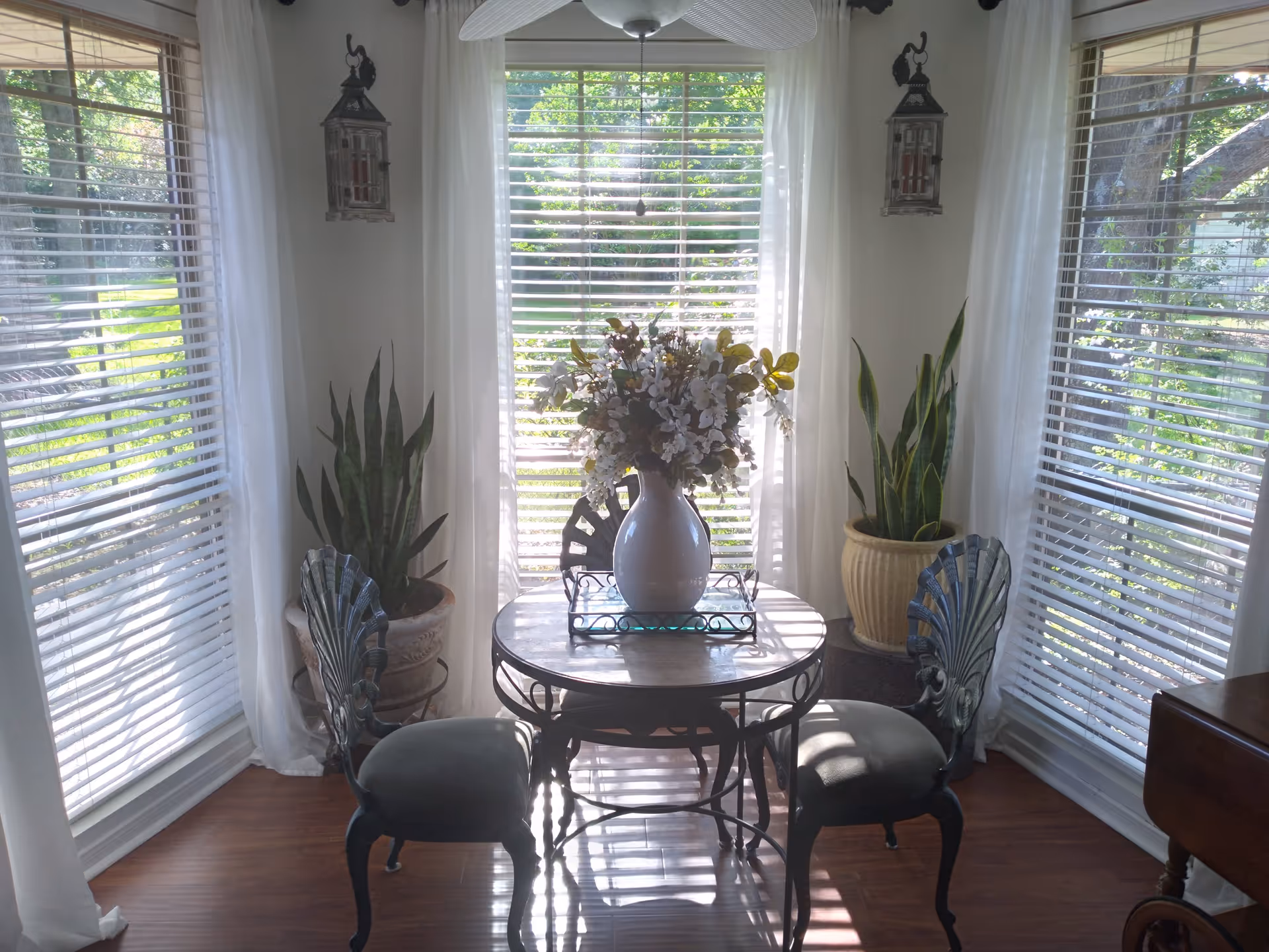 A small dining area with a round table and two ornate chairs. On the table is a white vase filled with a bouquet of flowers. The room has large windows with white blinds and sheer white curtains, allowing sunlight to stream in. There are two potted snake plants on either side of the table, and two decorative lanterns hanging on the walls. The floor is wooden.
