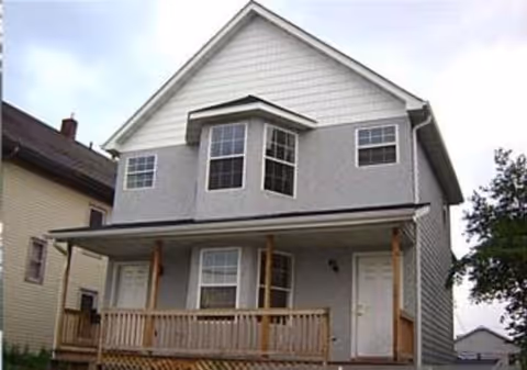 Front exterior view of a two-story gray house with white trim, featuring a small wooden porch with railings and two white doors. The house has multiple windows including a bay window on the second floor.