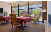 A bright dining room with tables covered in red tablecloths, surrounded by wooden chairs. Large floor-to-ceiling windows reveal a scenic view of mountains and greenery outside. The room has a cozy and inviting atmosphere with a chandelier overhead and a television mounted on the wall.