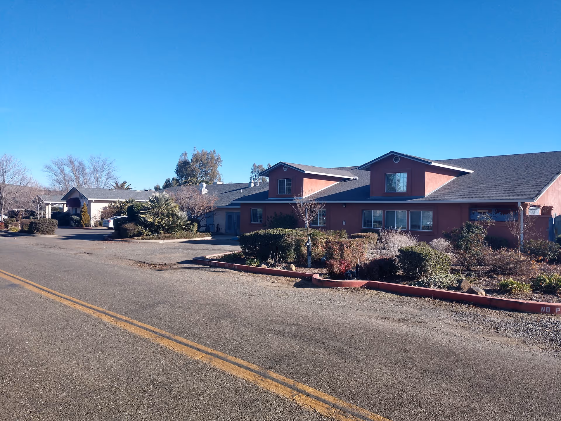 Exterior view of a single-story building with a reddish facade and multiple windows, surrounded by shrubs and small trees under a clear blue sky.