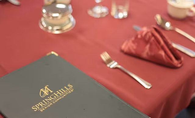 Close-up of a dining table set with a red tablecloth, silverware, a folded red napkin, a cup, and a green menu folder with the text 'Spring Hills Senior Communities' in gold lettering.