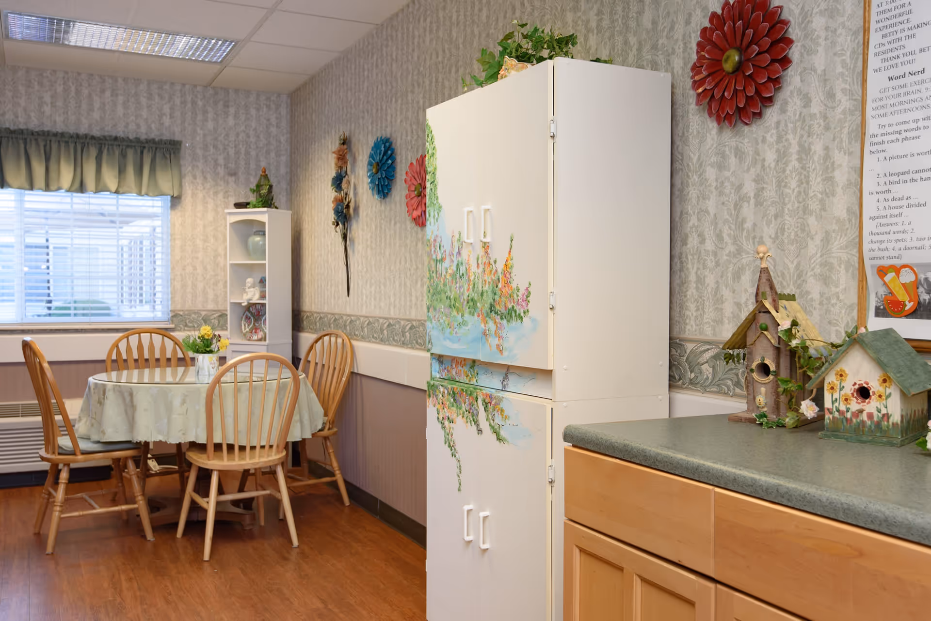 A cozy dining area with a round table covered with a light-colored tablecloth and surrounded by four wooden chairs. A window with green valance lets in natural light. The walls are decorated with floral wallpaper and colorful flower wall hangings. A white cabinet with a painted floral design stands against the wall, next to a countertop with two decorative birdhouses and some greenery.