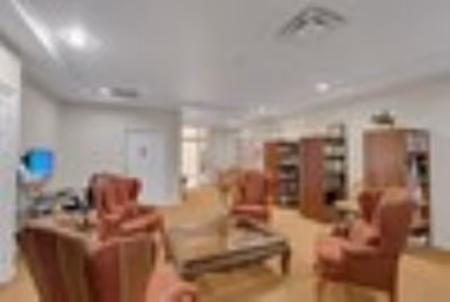 A cozy living room area with several red upholstered armchairs arranged around a wooden coffee table. The room has beige walls, carpeted flooring, and built-in bookshelves along the back wall. There is a computer station with a monitor on the left side near a window.