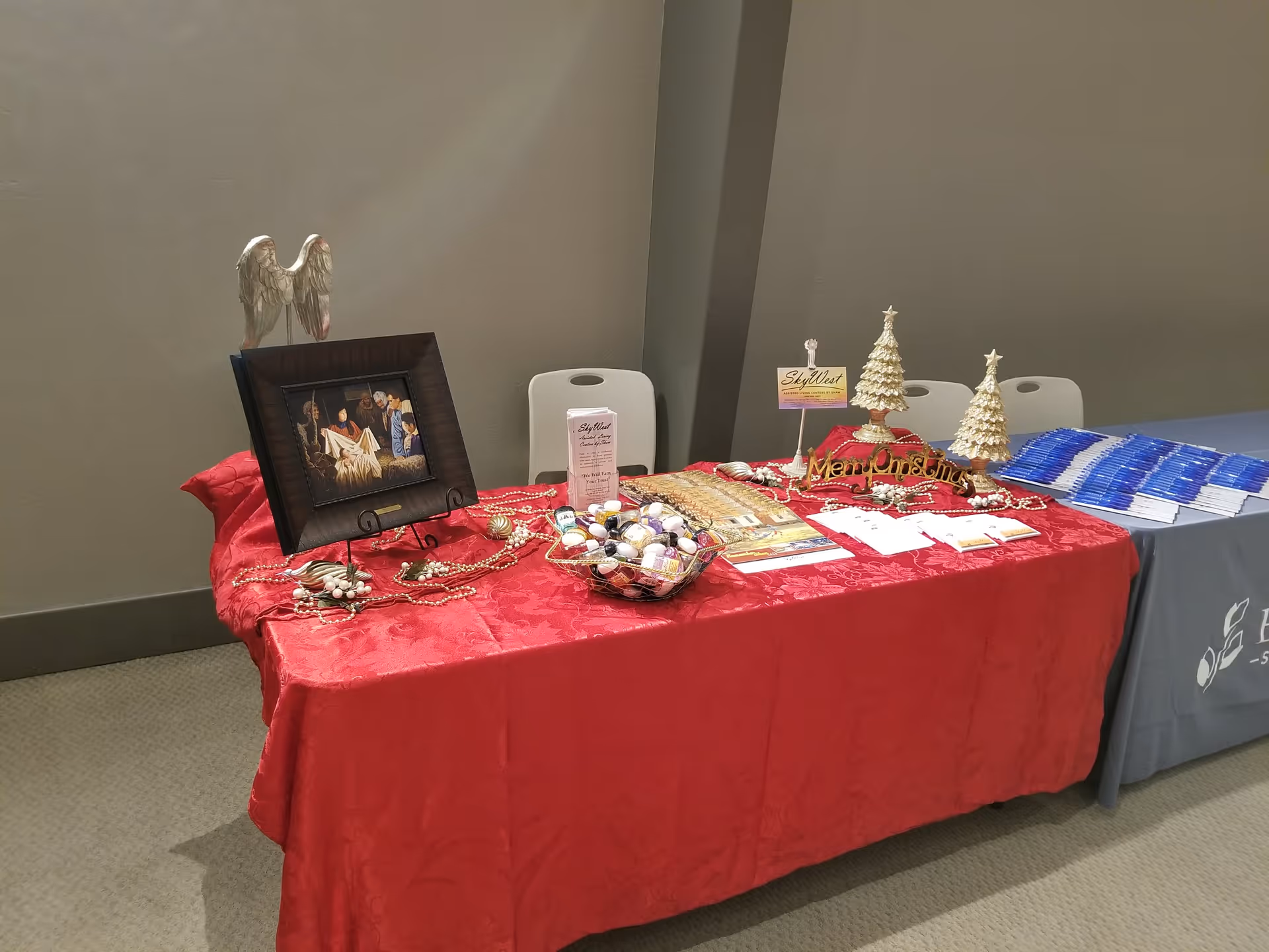 A table covered with a red tablecloth displaying Christmas decorations including two small white Christmas trees, a framed religious picture, a bowl of ornaments, and informational brochures. The table is set up indoors against a plain wall with two white chairs behind it.