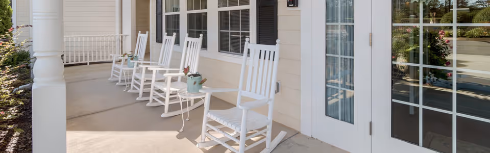 A covered outdoor porch area with several white wooden rocking chairs and small white tables, some with decorative watering cans. The porch has beige siding, white trim, and large windows with black shutters.