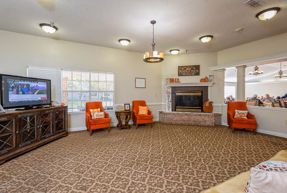 A cozy living room area in Sabine Place featuring a patterned carpet, three orange armchairs with decorative pillows, a wooden TV stand with a flat-screen TV, and a brick fireplace decorated with fall-themed items including pumpkins and a sign that reads 'Pumpkin Patch'. A window with blinds lets in natural light, and an adjacent room with people sitting at tables is visible through a large interior window.