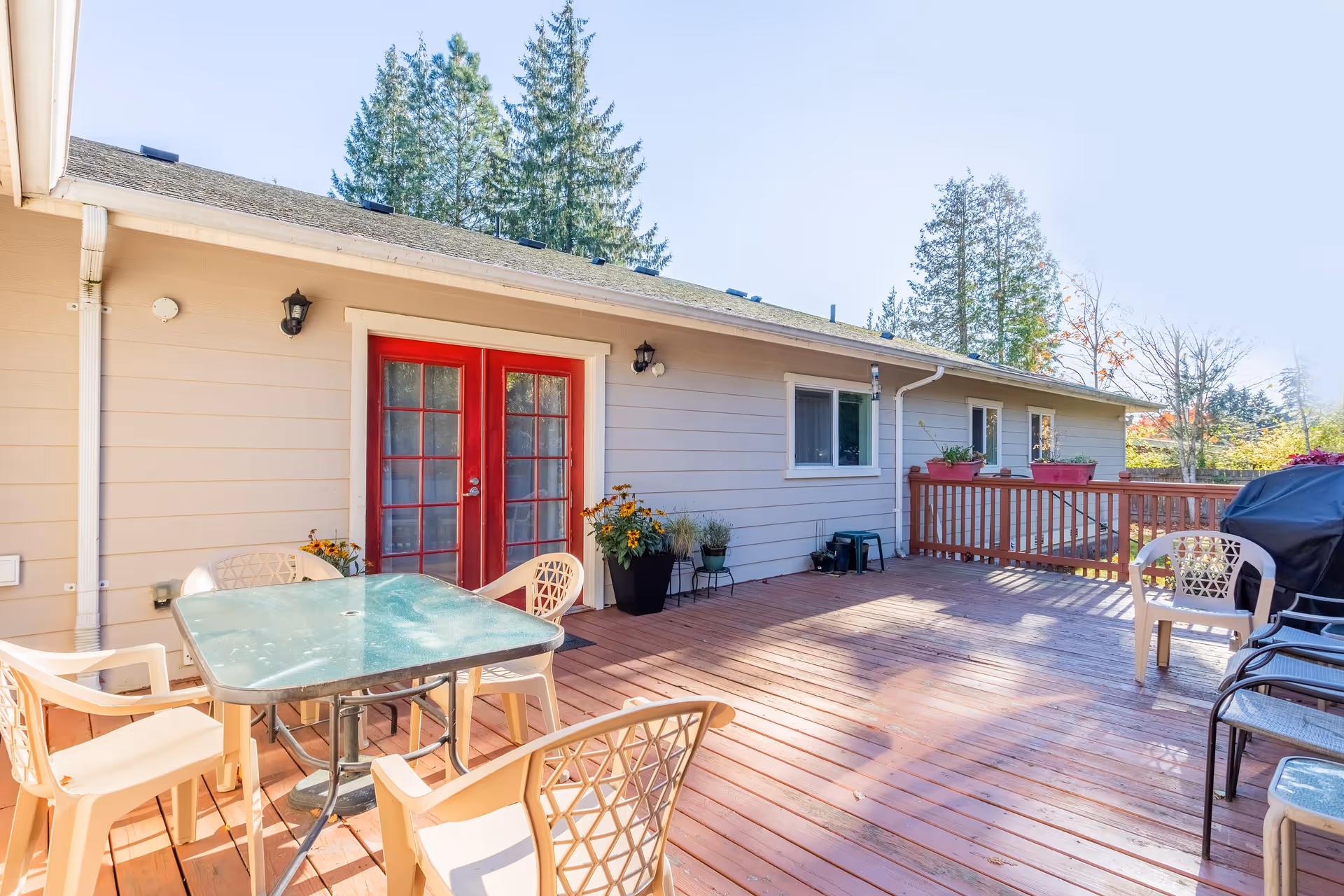 Outdoor wooden deck area with a glass-top table surrounded by plastic chairs, a barbecue grill, potted plants, and red double doors leading into a beige building. Tall trees are visible in the background under a clear blue sky.