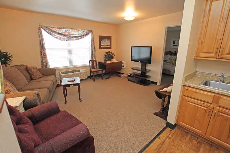 A cozy senior living room with a sofa, armchair, TV on a stand, small tables, and a kitchenette area visible to the right.