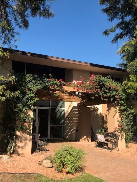Outdoor seating area at Ocotillo Place featuring two benches under a pergola adorned with flowering vines. The building exterior is beige with large windows, surrounded by trees and greenery under a clear blue sky.