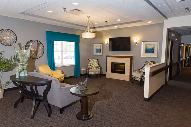 A cozy common area in a senior living facility featuring a fireplace with a mounted TV above it, two patterned armchairs, a yellow armchair near a window with teal curtains, a gray armchair, a round side table with a cat figurine, a black console table with a decorative sculpture, and wall art. The space has carpeted flooring and a hallway leading to other rooms.