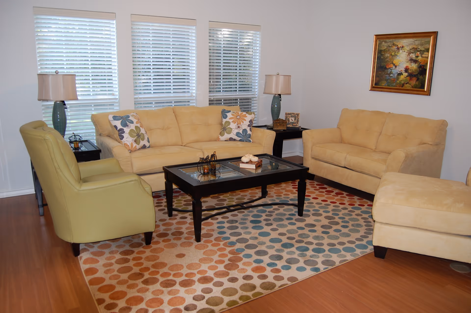 A cozy living room with two beige sofas, a green armchair, and a matching beige ottoman arranged around a black coffee table with glass panels. The floor has a colorful polka dot patterned rug, and there are two table lamps on side tables beside the sofas. Three windows with white blinds are in the background, and a framed painting hangs on the wall.