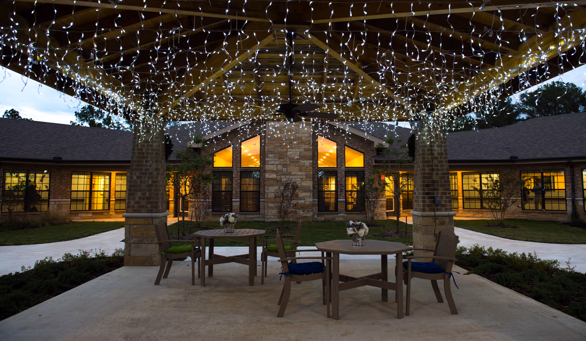 Covered outdoor patio courtyard with hanging string lights, tables and chairs in front of a lit senior living building at dusk.