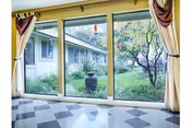 View through large glass sliding doors showing a garden area with trees, shrubs, and a large decorative urn outside. The interior floor has a checkered tile pattern, and cream-colored curtains with maroon accents frame the doors.