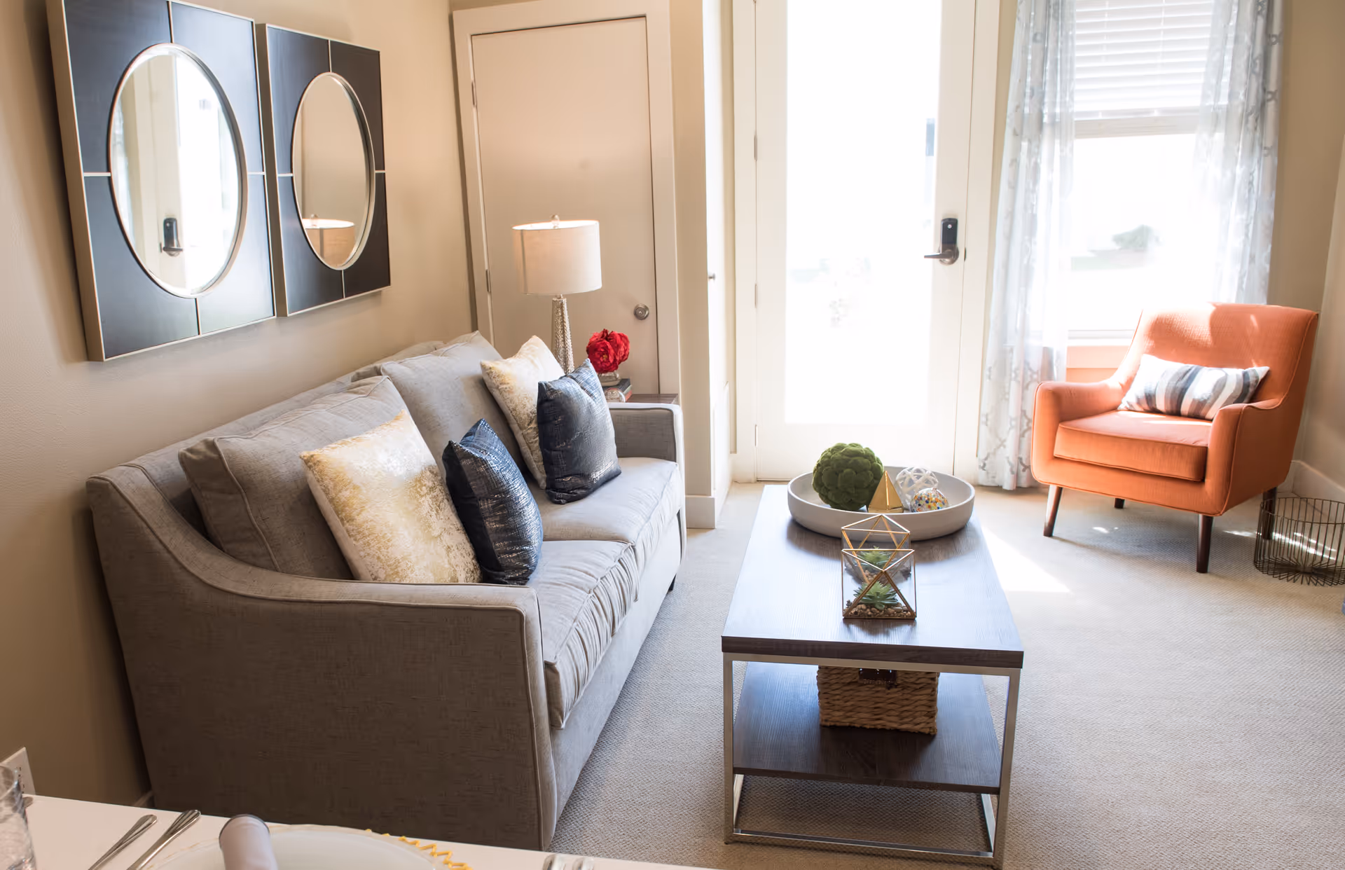 A bright and cozy living room with a gray sofa adorned with decorative pillows, a wooden coffee table with decorative items, an orange armchair with a striped pillow, two round mirrors on the wall, a table lamp, and a door with a window letting in natural light.