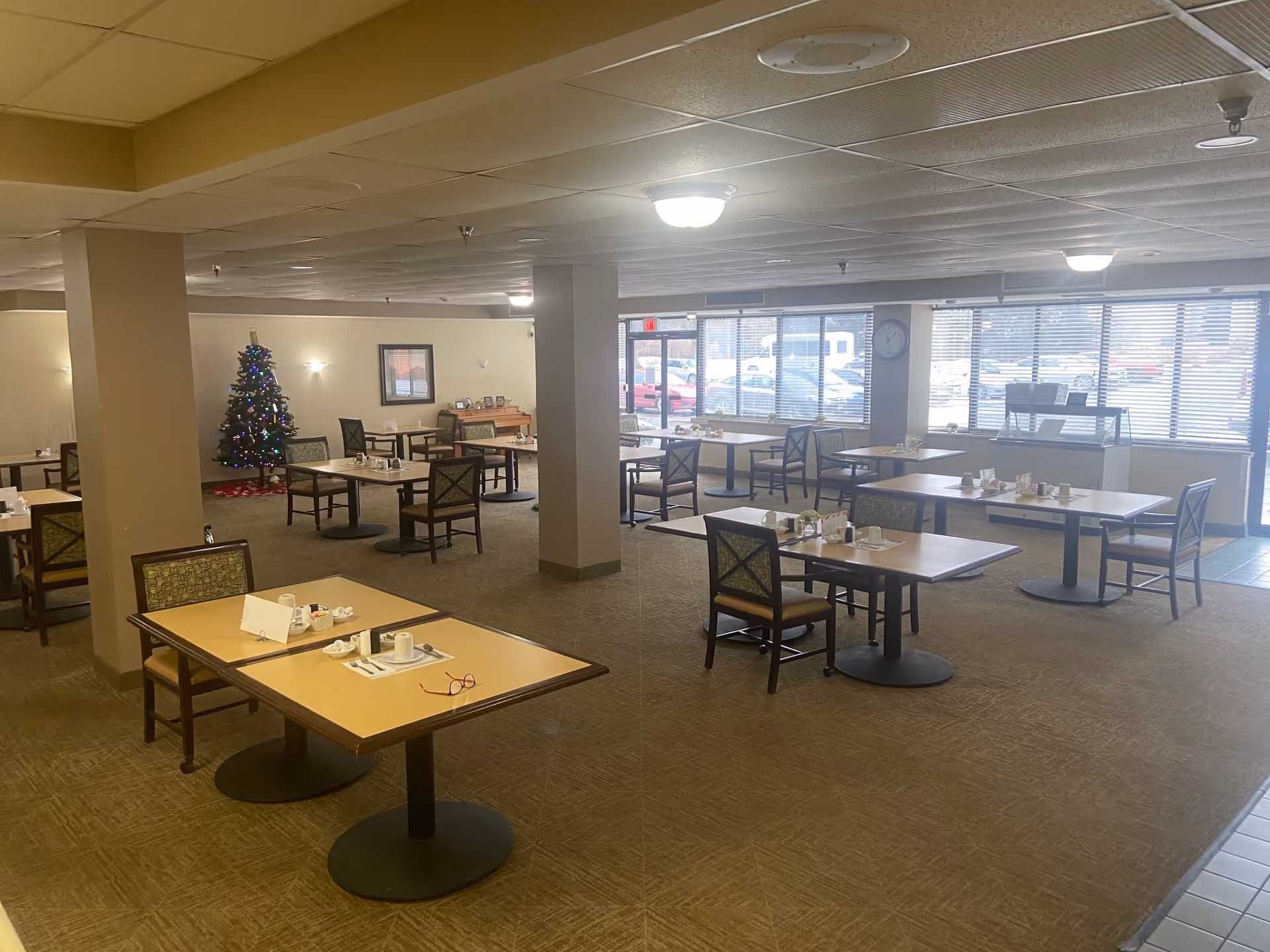 Dining room with several tables and chairs set for meals, a decorated Christmas tree in the corner, and large windows along the front.