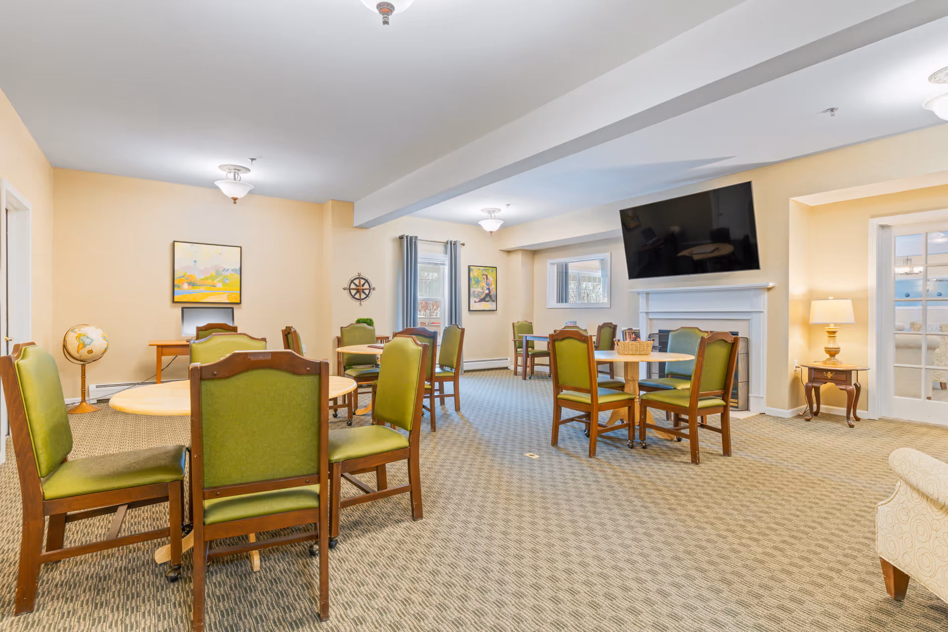 Bright communal room with several round tables and green-upholstered chairs, a wall-mounted TV above a fireplace, and framed artwork and lamps.