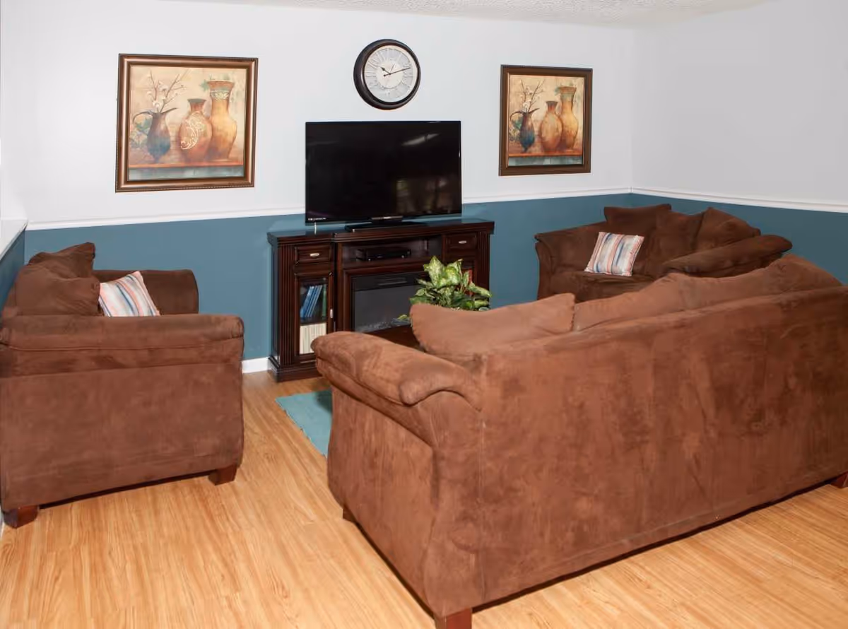 Small living room with brown sofas arranged around a TV on a wooden stand, framed artwork and a clock on the wall.