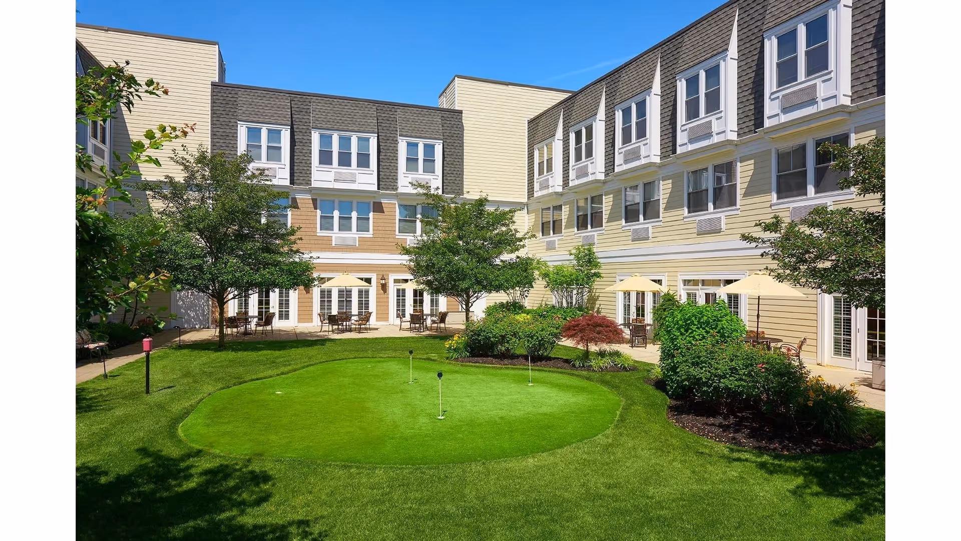 Sunlit courtyard at an assisted living facility with a putting green, patio tables and umbrellas, landscaping, and a surrounding multi-story building.