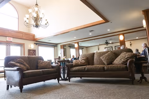 A spacious living room area in a senior living facility with two large brown leather sofas with patterned cushions, a chandelier hanging from a high ceiling, large windows letting in natural light, and several elderly people seated at tables in the background.