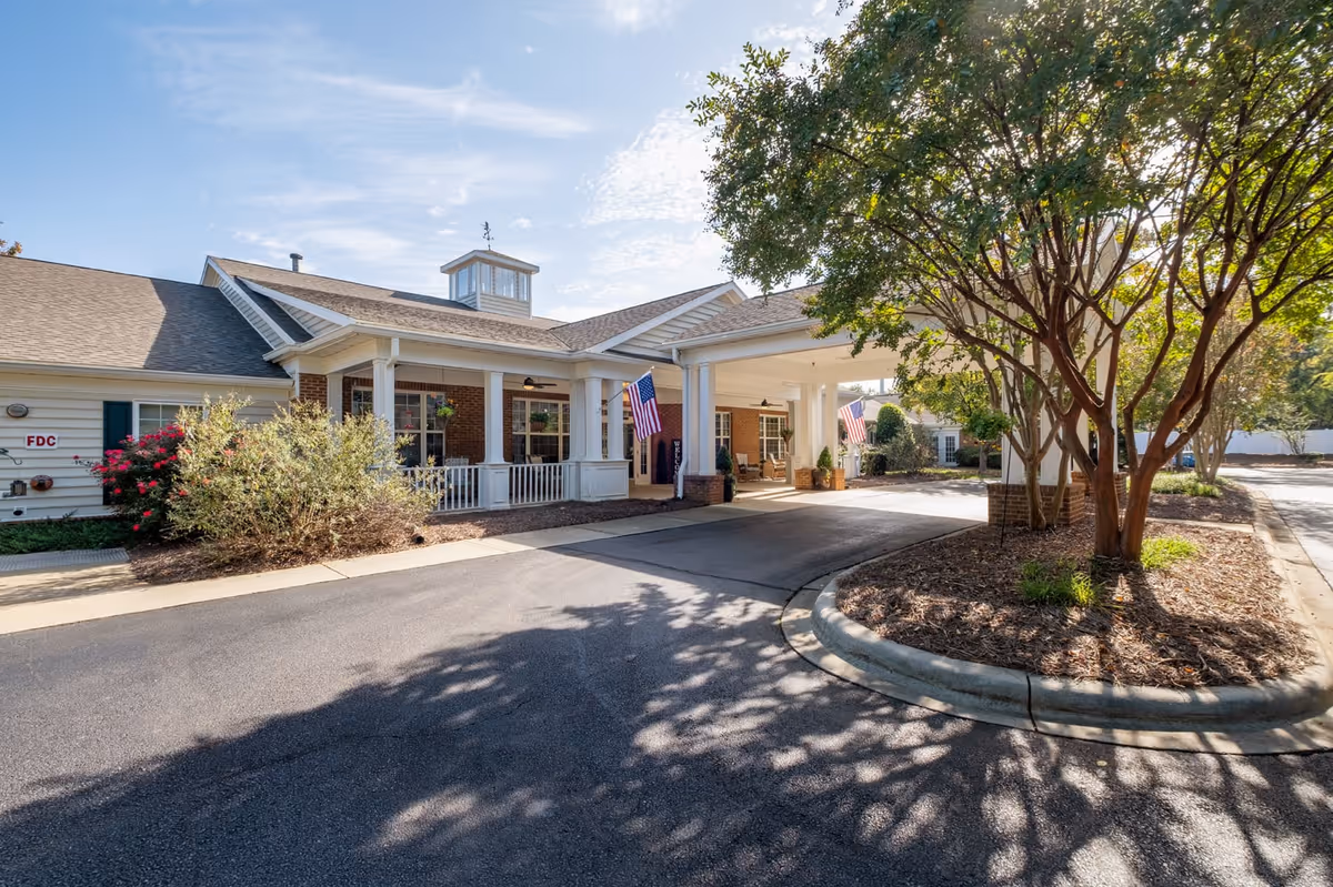 Exterior view of Chandler Place Assisted Living and Memory Care facility showing the entrance with a covered driveway, American flags, brick and siding walls, and landscaped trees and bushes under a clear sky.