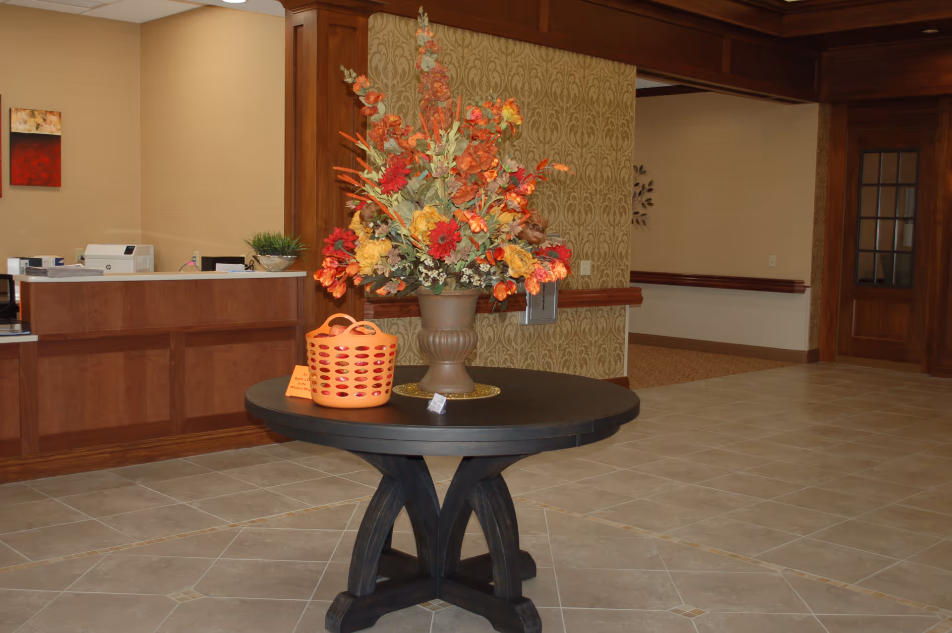 Interior view of a senior living facility lobby area with a round black table in the center holding a large vase of colorful artificial flowers and an orange basket. Behind the table is a wooden reception desk with office equipment and a beige wall with framed artwork. The floor is tiled and there is a wooden door with glass panels on the right side.