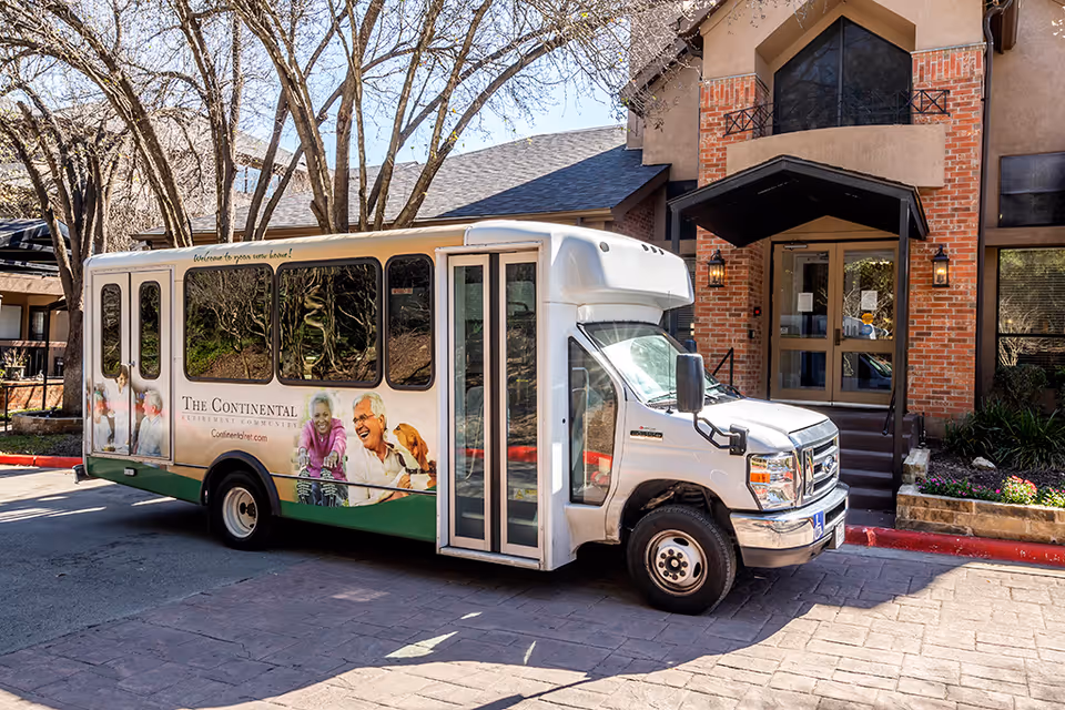 A white shuttle bus with images of smiling elderly people and a dog parked in front of the entrance to a brick and stucco building at The Continental Retirement Community. The bus has large windows and the text 'The Continental Retirement Community' along with the website 'Continentalret.com' on its side.