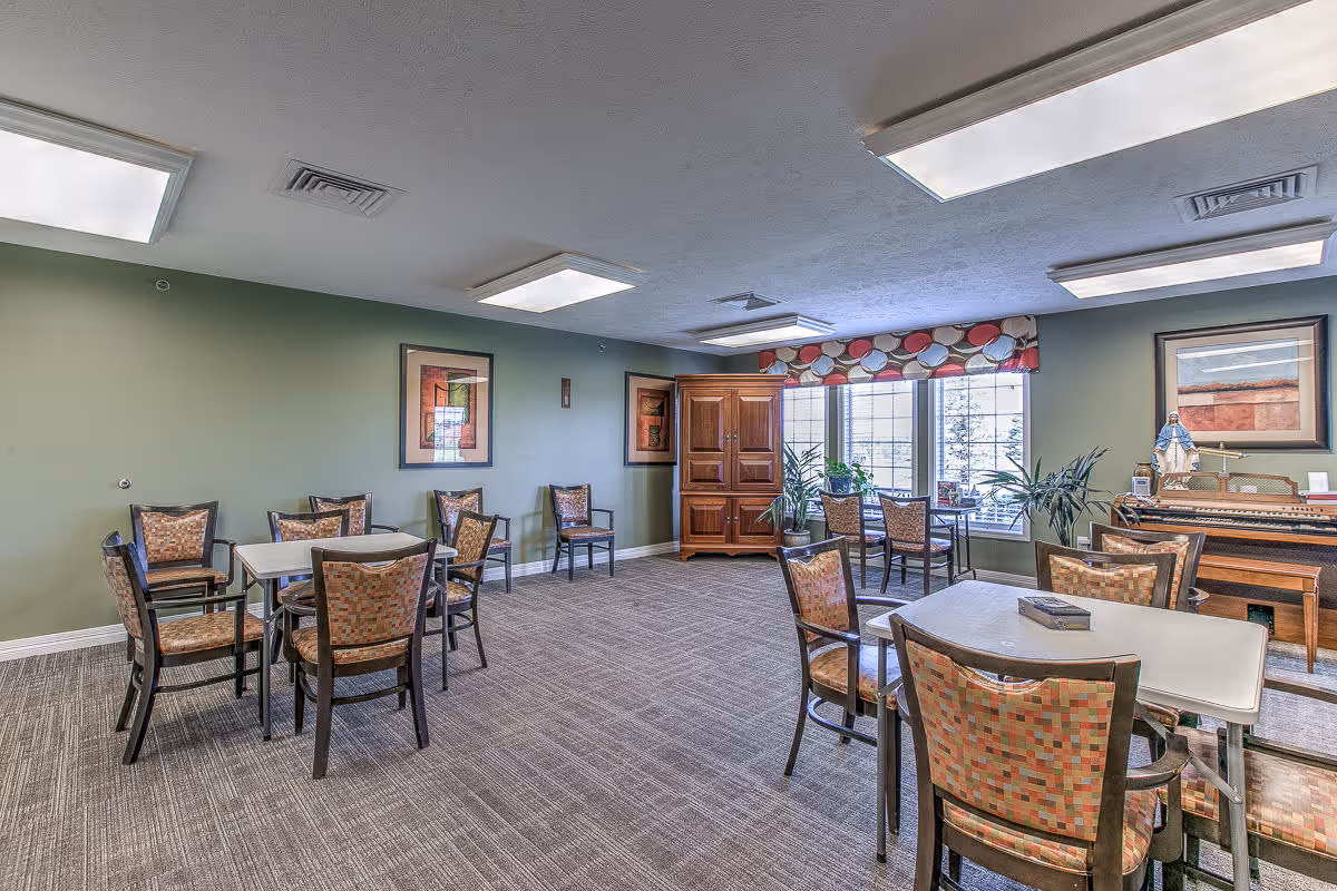 A spacious room with multiple tables and chairs arranged for group seating. The walls are painted green and decorated with framed artwork. Large windows with patterned valances allow natural light to enter. There is a wooden cabinet and a piano against the far wall, along with some potted plants.