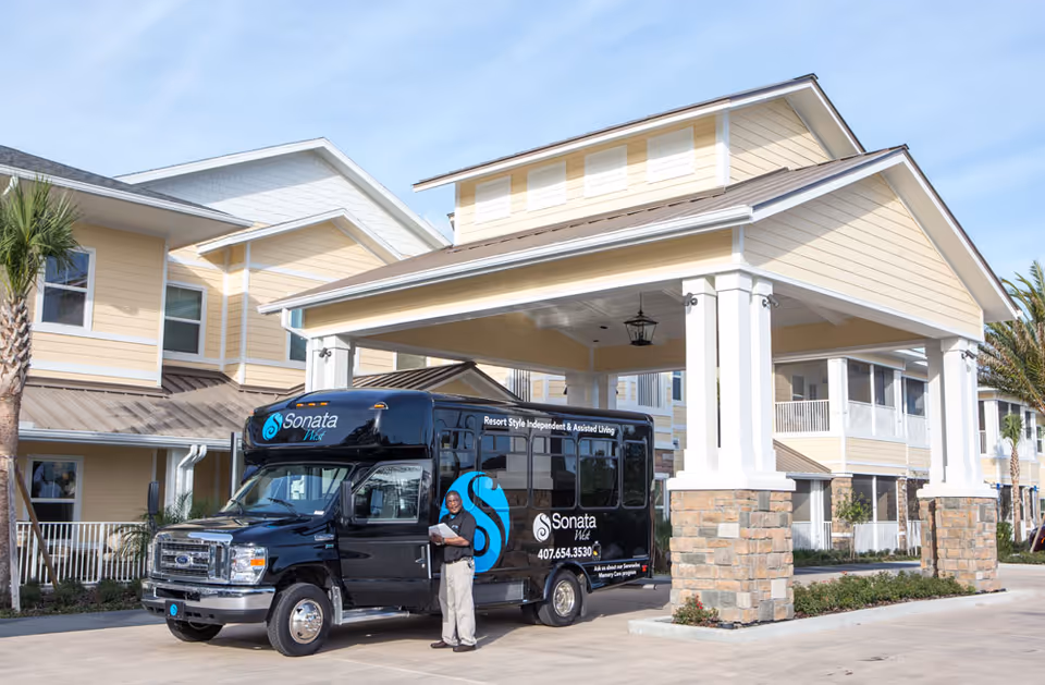 Porte-cochere entrance of a senior living building with a black Sonata shuttle bus parked beneath and a staff member standing beside it.