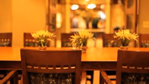 A warmly lit dining room with a long wooden table surrounded by wooden chairs. Small flower arrangements are placed on the table, and the background shows a softly blurred interior space with warm lighting.