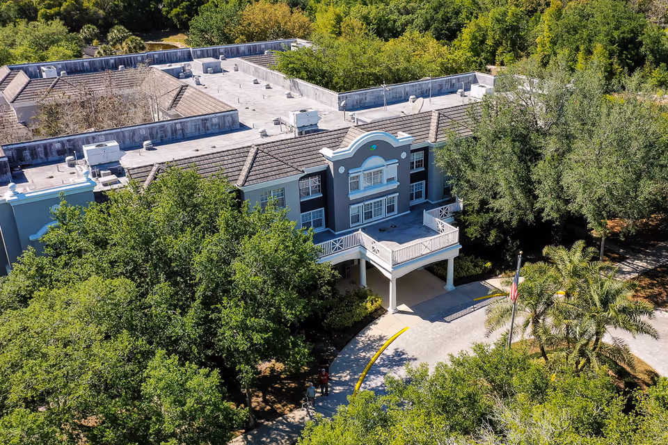 Aerial view of Tampa Gardens Senior Living facility showing the front entrance of a multi-story building surrounded by lush green trees and landscaping, with a curved driveway and an American flag near the entrance.