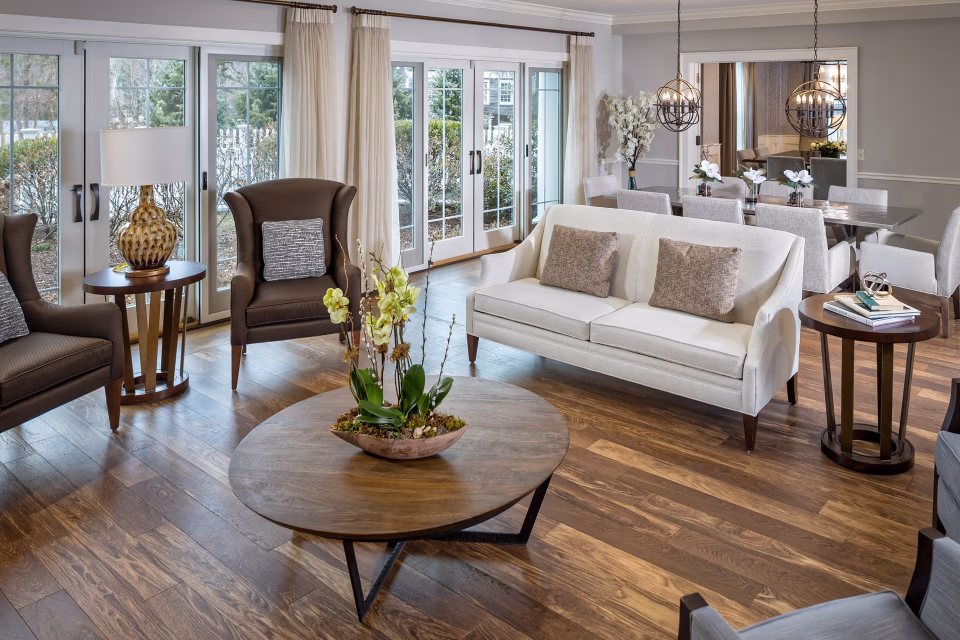 A bright and elegant living room area with wooden flooring, featuring a white sofa with two brown cushions, two dark brown armchairs with gray pillows, a round wooden coffee table with a floral centerpiece, and side tables with a lamp and books. Large glass doors with white curtains open to an outdoor view. In the background, there is a dining area with a table, chairs, decorative flowers, and two spherical pendant lights.
