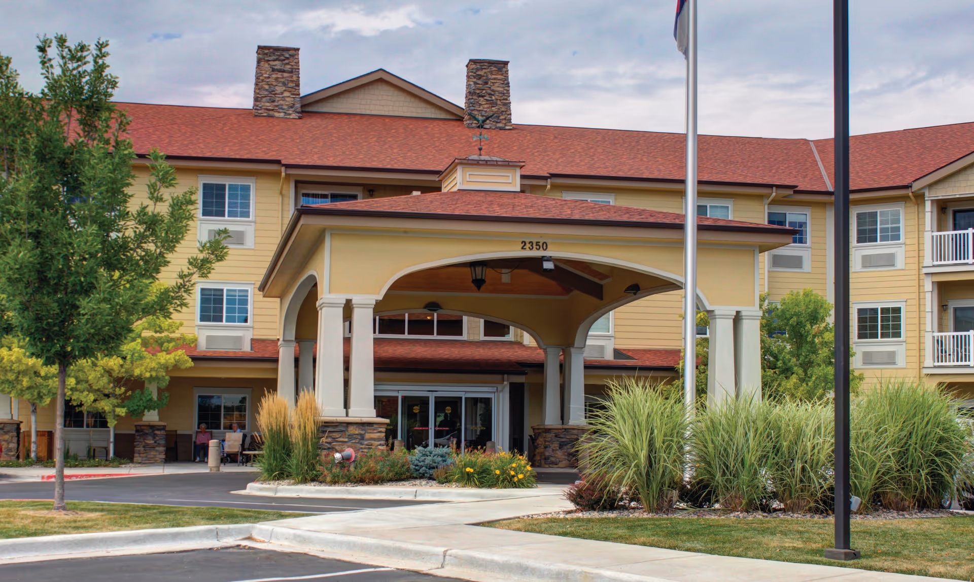 Front exterior view of Rigden Farm Senior Living building with a covered entrance, stone pillars, and landscaped greenery including trees and bushes. The building has a red roof and yellow siding with multiple windows.
