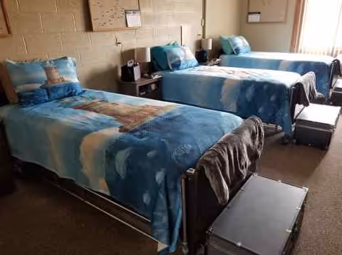 A senior living facility bedroom with three single beds arranged side by side. Each bed has blue bedding with lighthouse and ocean designs, and a pillow with a lighthouse image. There are small nightstands between the beds with lamps and a clock. The room has a window with vertical blinds letting in natural light.