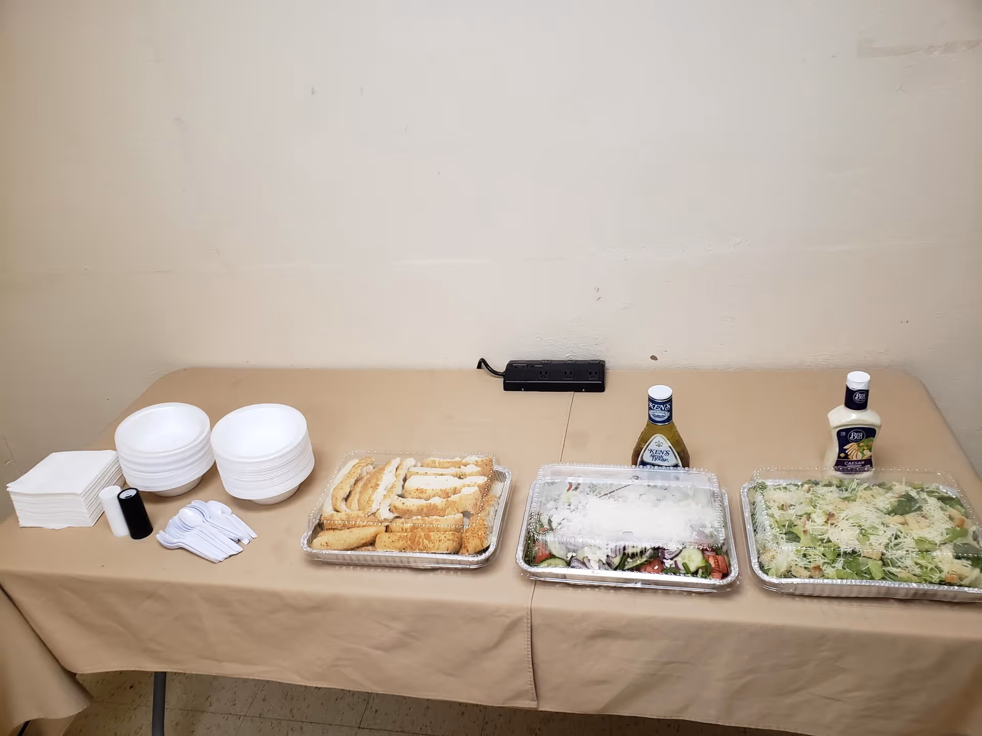A table covered with a beige tablecloth displaying disposable plates, bowls, napkins, plastic spoons, salt and pepper shakers, a tray of breadsticks, and two trays of salad with bottles of salad dressing on a plain beige wall background.