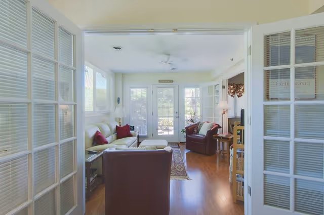 A bright living room with wooden floors, a white sofa with red pillows, a brown armchair, a small table with a lamp, and large windows and glass doors letting in natural light. The room is viewed through open French doors with glass panes.