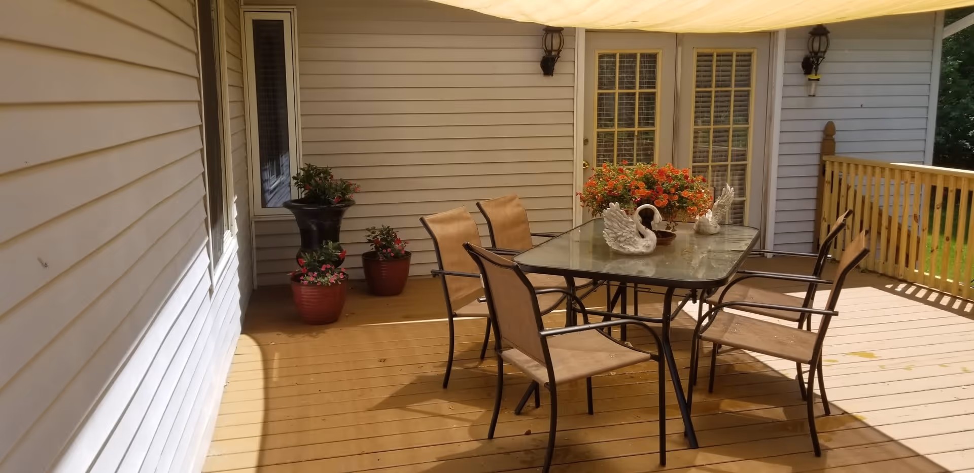 Outdoor patio area with a glass-top table surrounded by six brown mesh chairs. The table has two white swan figurines and a pot of orange flowers. The patio has wooden flooring and beige siding walls with two wall-mounted lantern lights. There are three potted plants in the corner near a window and a set of French doors.