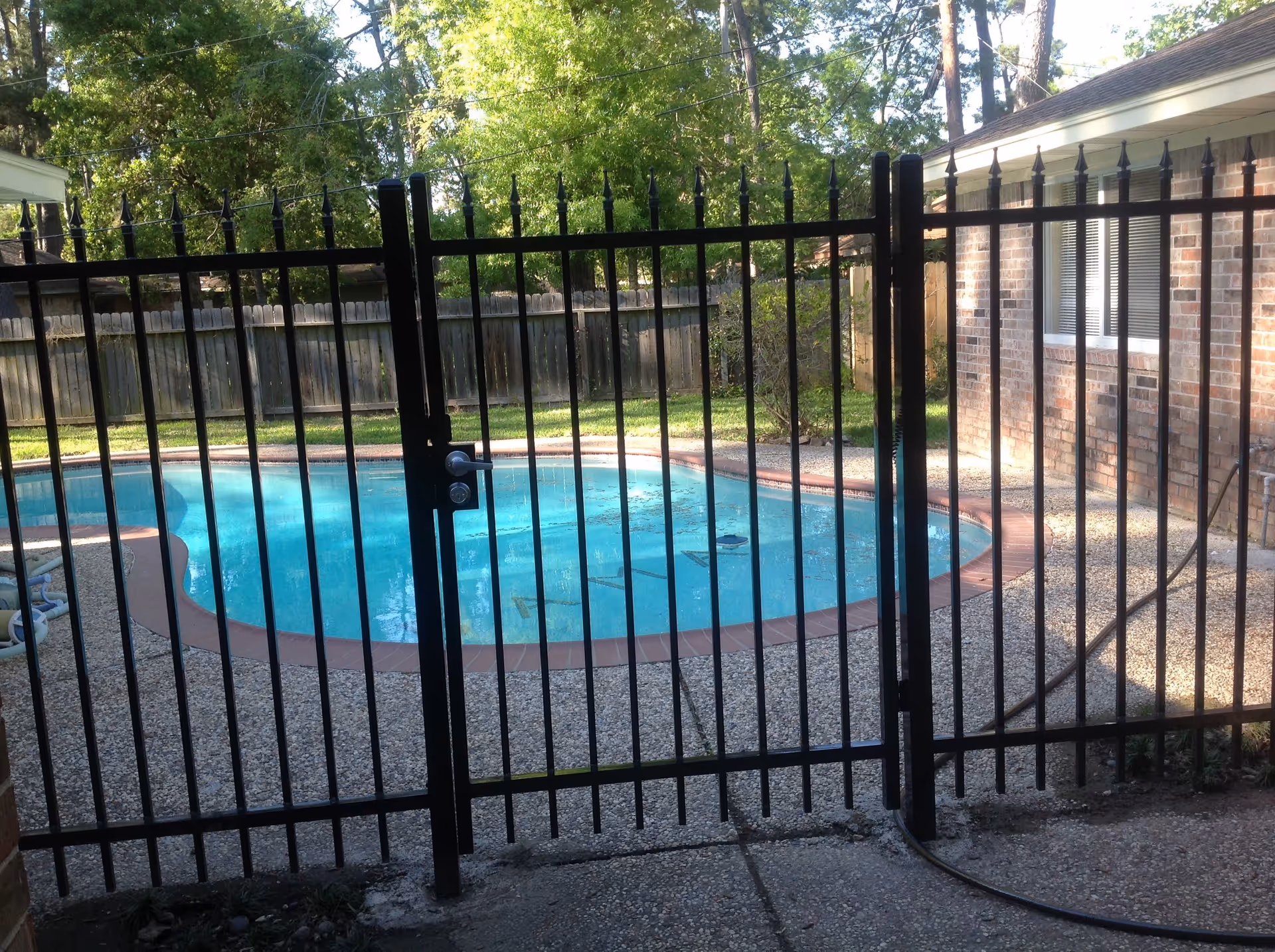 View of a fenced swimming pool in a backyard with a brick house on the right and trees in the background.
