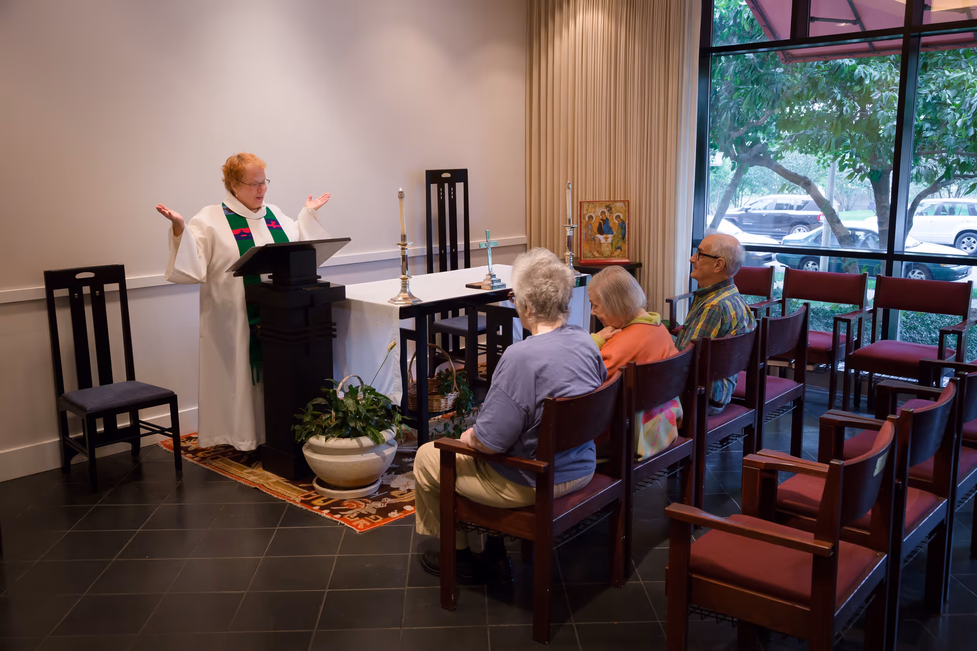 A small group of elderly people seated in wooden chairs with red cushions in a room with large windows. A clergy member in white robes with a green stole stands at a lectern with arms raised, leading a service. The room has a table covered with a white cloth, candles, a cross, and a religious icon. Outside the window, trees and parked cars are visible.