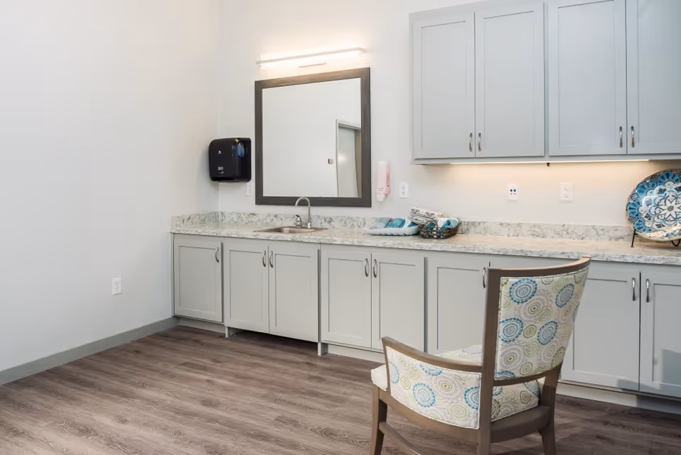 A bright interior room with a long countertop and sink, upper and lower cabinets, a mirror, and a patterned upholstered chair on wood-look flooring.
