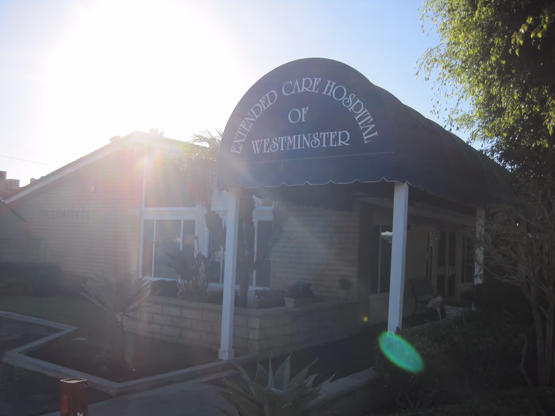 Exterior view of the entrance to Extended Care Hospital of Westminster with a black awning displaying the facility name. The building is partially obscured by sunlight and surrounded by plants and trees.