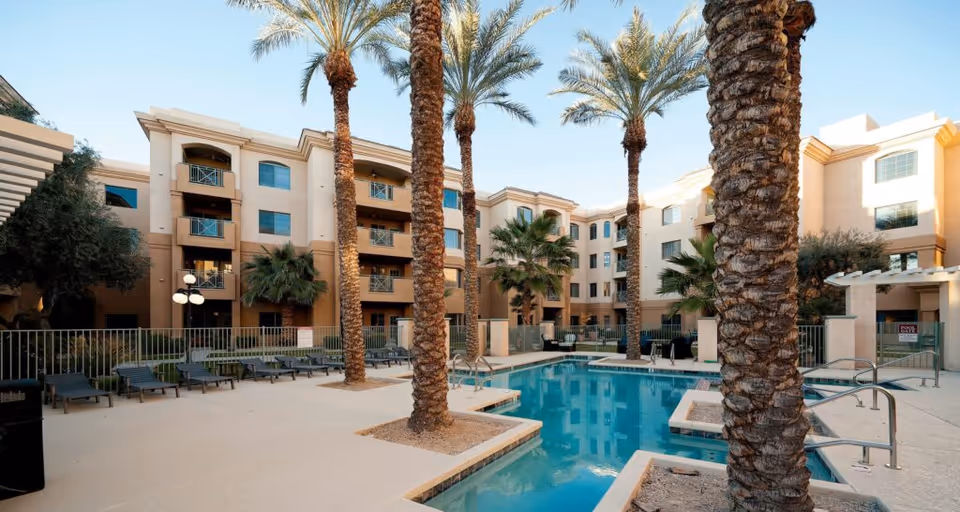 Courtyard with a swimming pool, palm trees, lounge chairs, and a multi-story residential building in the background.