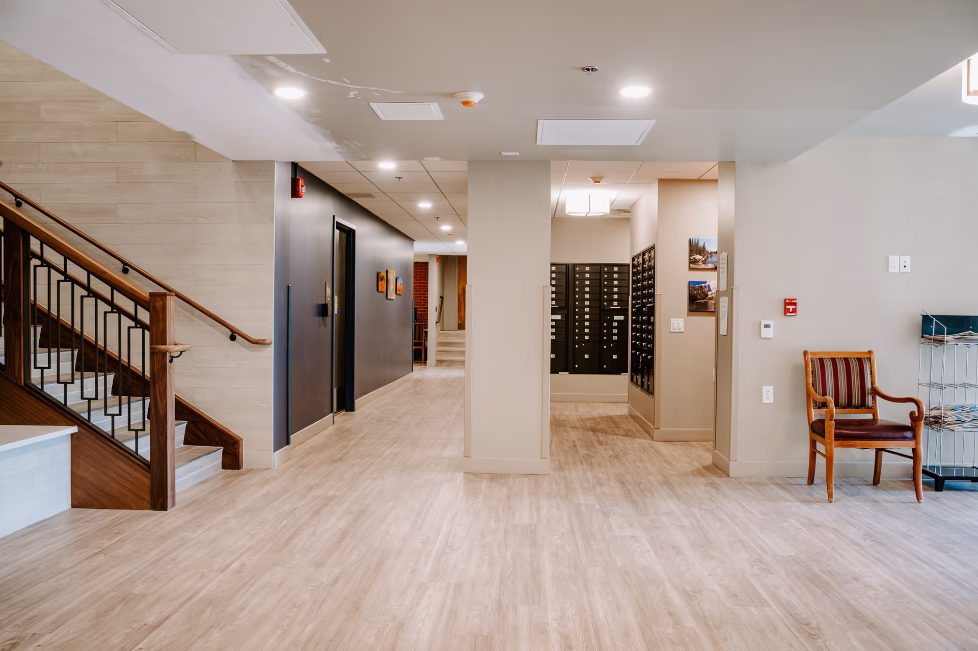 Interior hallway of a senior living facility with light wood flooring, beige walls, a staircase with wooden handrails on the left, a row of mailboxes in the center background, a wooden chair with striped upholstery on the right, and a metal rack with magazines.