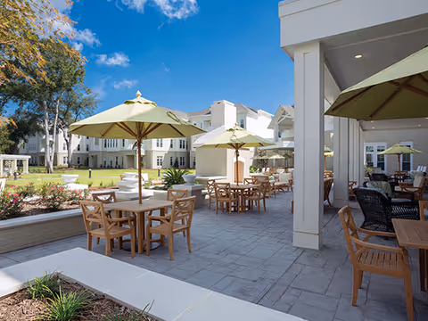 Outdoor patio with wooden tables, chairs and green umbrellas on a paved terrace beside a multi-story senior living building under a bright blue sky.
