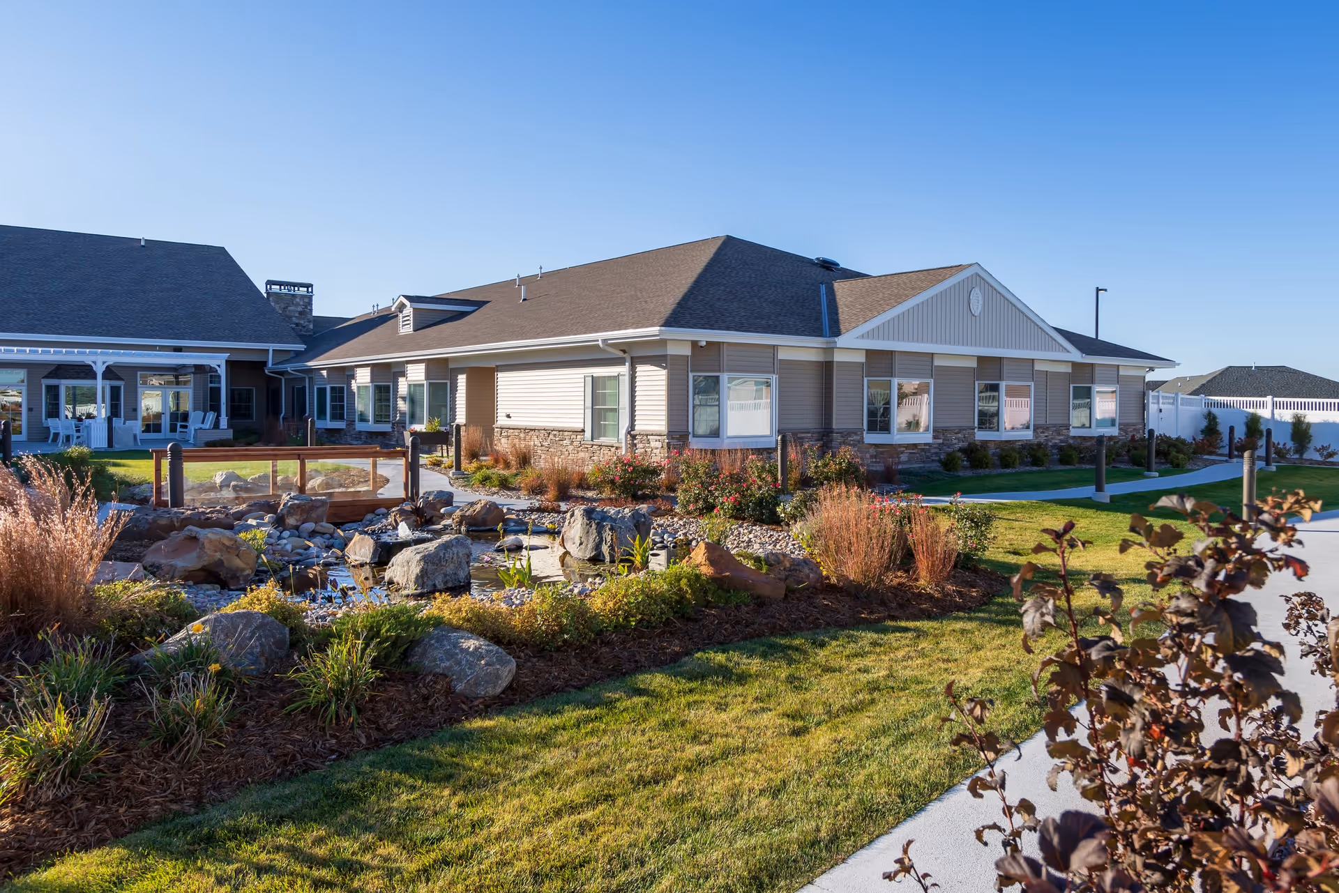 Exterior view of CountryHouse Residence, a single-story building with beige siding and stone accents, surrounded by a landscaped garden with rocks, plants, and a small pond under a clear blue sky.