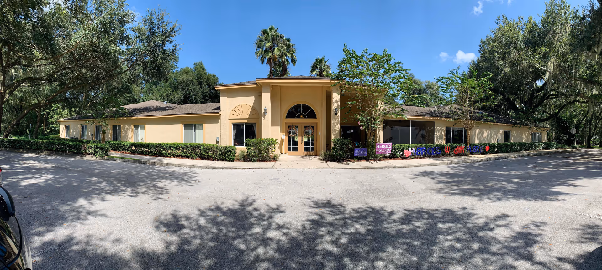 Exterior front view of a single-story senior living facility building with a beige facade, large entrance with double doors, surrounded by trees and bushes under a clear blue sky.