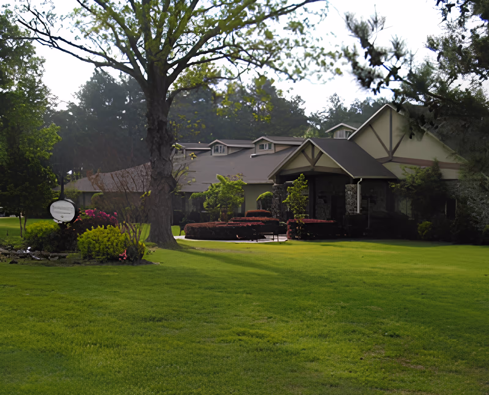 A large single-story building with a peaked roof and stone accents, surrounded by a well-maintained green lawn and various trees and shrubs. The scene is outdoors with natural light and a peaceful garden setting.