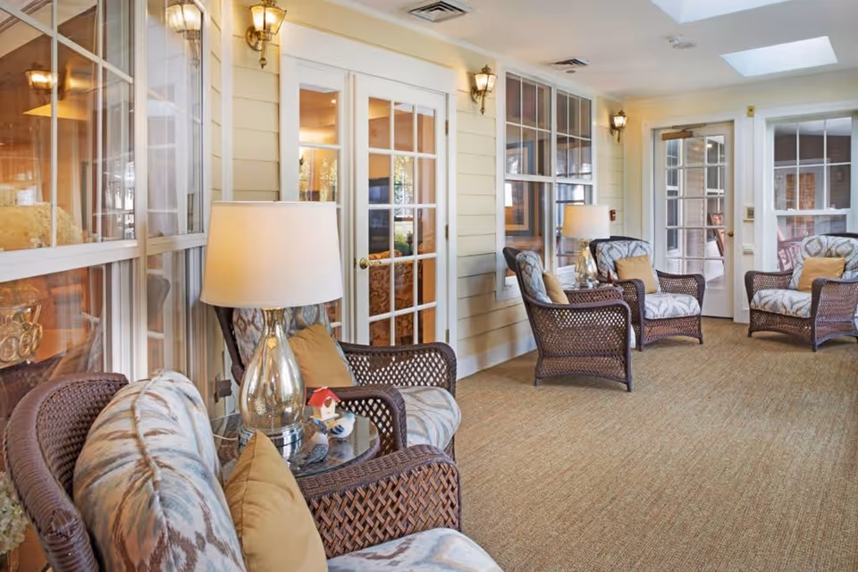 A bright and cozy indoor seating area with wicker armchairs featuring patterned cushions and beige pillows. There are glass-top side tables with decorative lamps and small ornaments. The walls are light-colored with multiple windows and French doors, and the floor is carpeted in a neutral tone. Wall-mounted lantern-style lights provide additional lighting.
