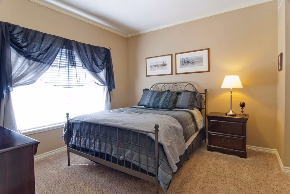 A cozy bedroom with a metal frame bed dressed in gray and blue bedding. There is a wooden nightstand with a lit lamp on the right side of the bed. Two framed pictures hang on the beige wall above the bed. A window with sheer dark blue curtains allows natural light to brighten the room. The floor is carpeted in a light beige color.